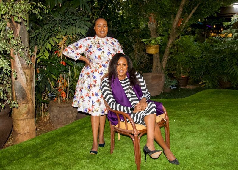 An image of two women posing for a picture on a green lawn. One is sitting on a chair while the other stands behind her